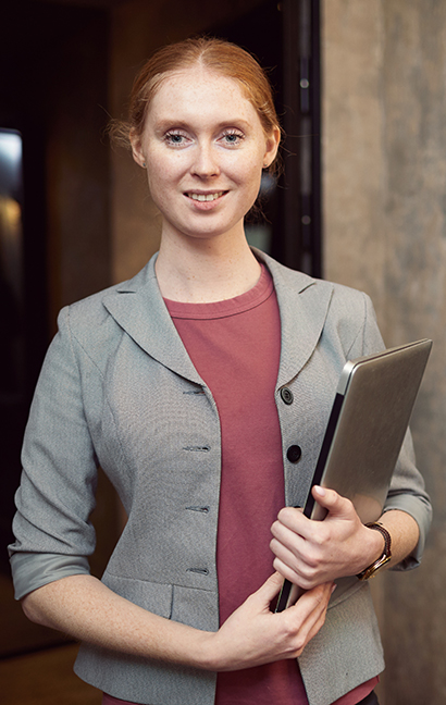 Portrait of young red haired manager holding laptop and looking at camera while working at office